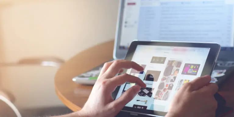 Person using a tablet to browse images, with an open laptop displaying email in the background on a wooden table.