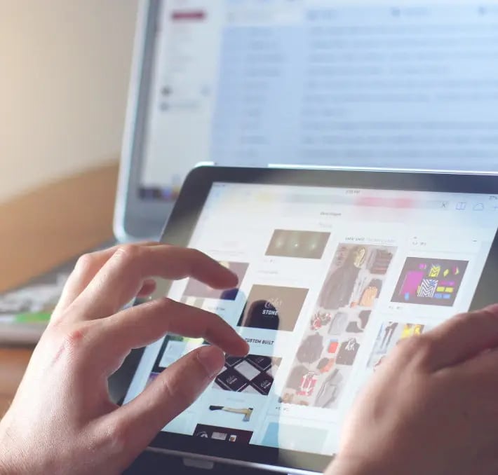 Person browsing an online store on a tablet with a laptop in the background displaying an email inbox.