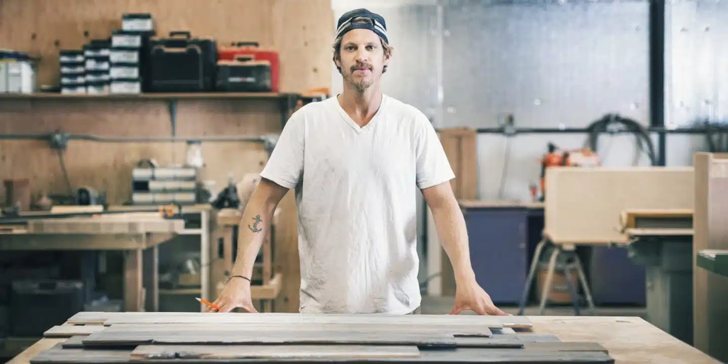 A man in a white t-shirt and backwards cap stands behind a workbench in a woodworking shop, with tools and materials in the background.
