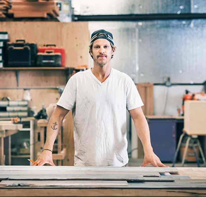 A man in a white t-shirt and backwards cap stands behind a workbench in a woodworking shop, with tools and equipment visible in the background.