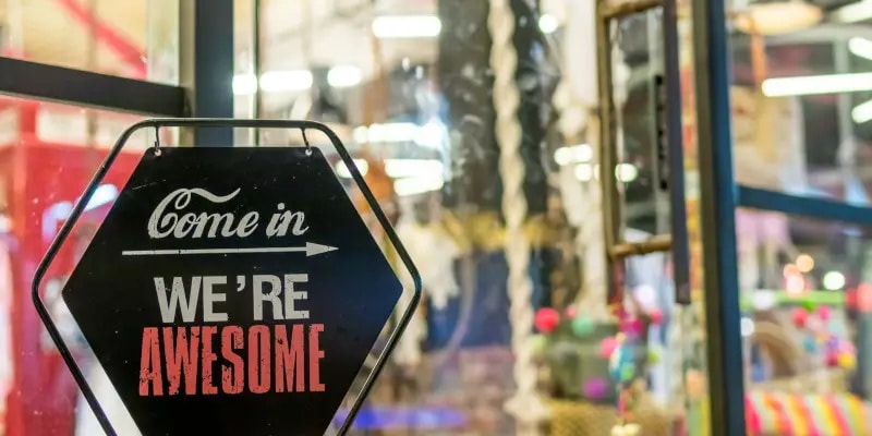 A black sign with white and red text reads, Come in, WERE AWESOME, hanging on a glass door with blurred lights and colors in the background.