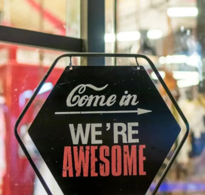 Hexagon-shaped sign on a glass door reads Come in, WERE AWESOME with an arrow pointing right; background shows an indoor setting.