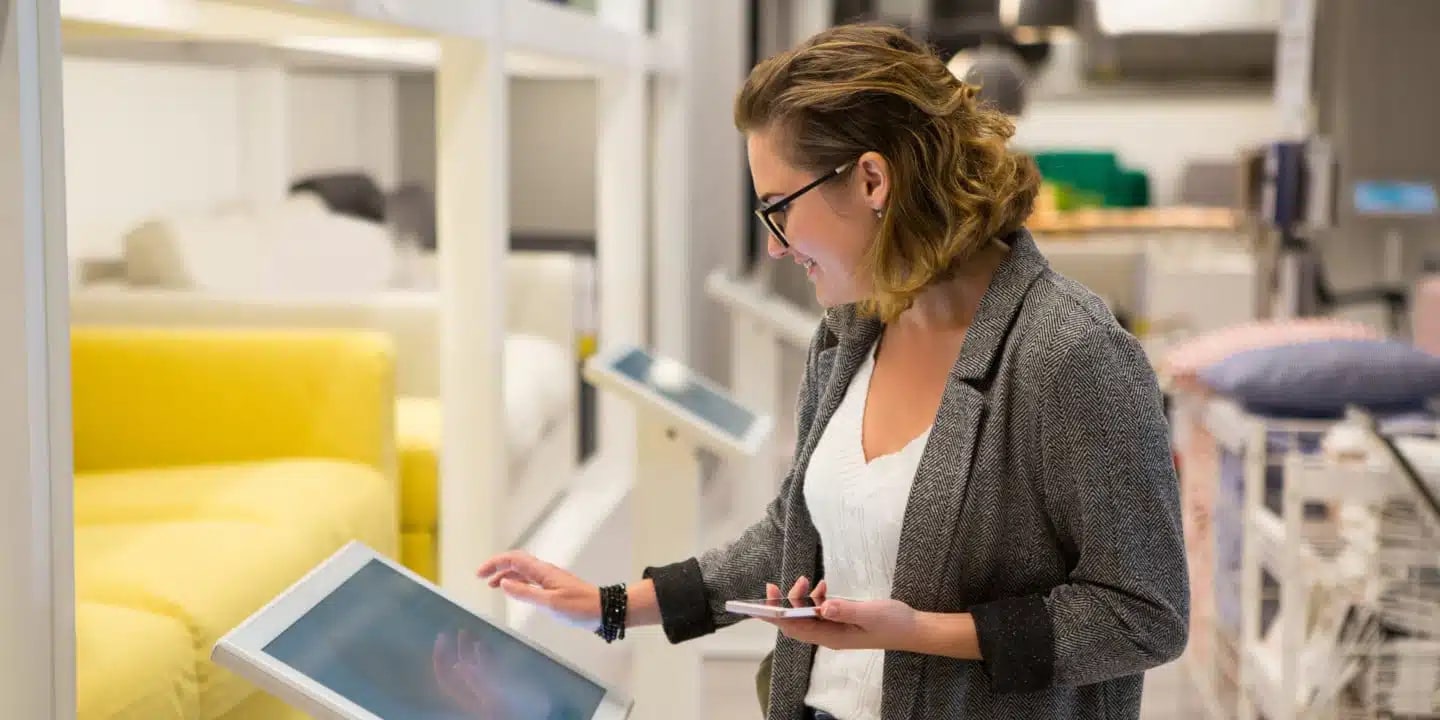 A woman stands indoors using a touchscreen kiosk while holding a smartphone, with furniture displays visible in the background.