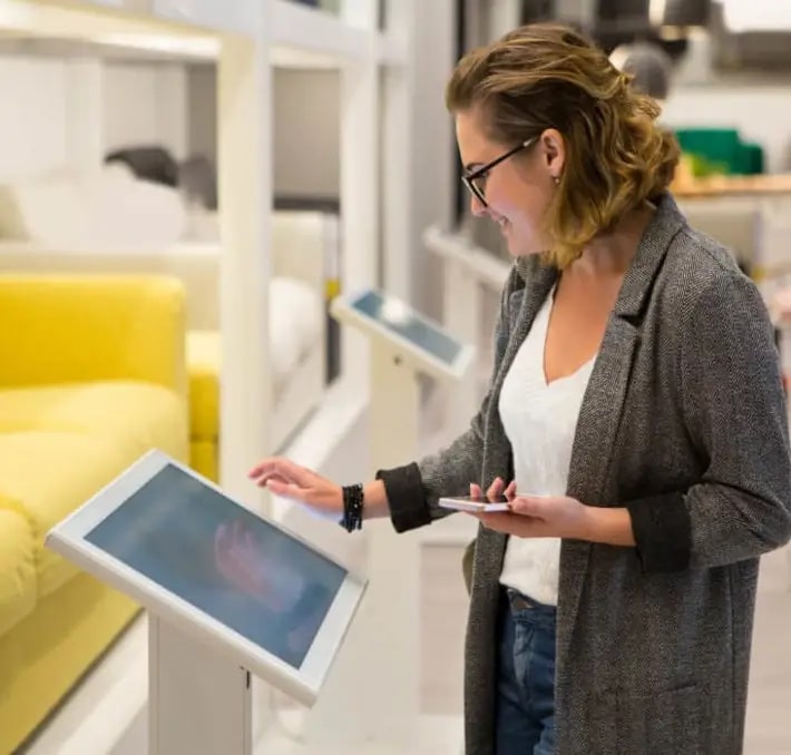 A woman uses a touchscreen kiosk in a modern retail store, holding her phone in one hand and interacting with the screen with the other.