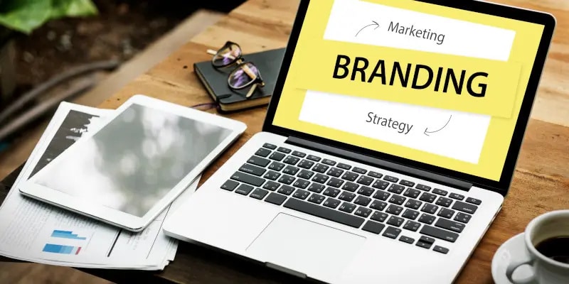 A laptop on a desk displays a branding concept with arrows between marketing and strategy. Nearby are papers, a pen, eyeglasses, and a smartphone.