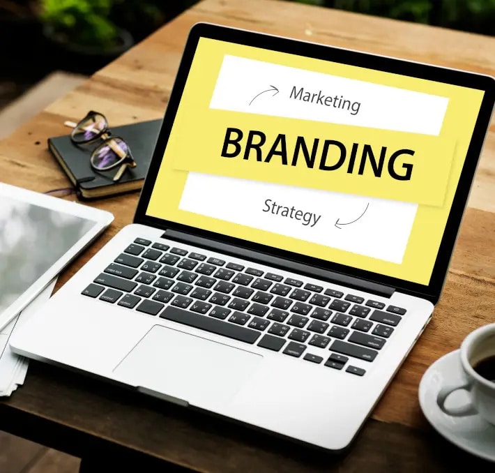 Open laptop on a wooden desk displays a slide with the word BRANDING and arrows pointing to Marketing and Strategy. Glasses, paper, and a cup are beside the laptop.