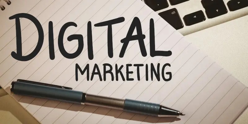 A notebook with DIGITAL MARKETING written on it, a pen lying on the notebook, and part of a laptop keyboard visible in the background.