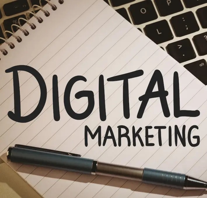 A notebook with DIGITAL MARKETING written on a page, a blue pen placed on the notebook, and part of a laptop keyboard visible in the background.
