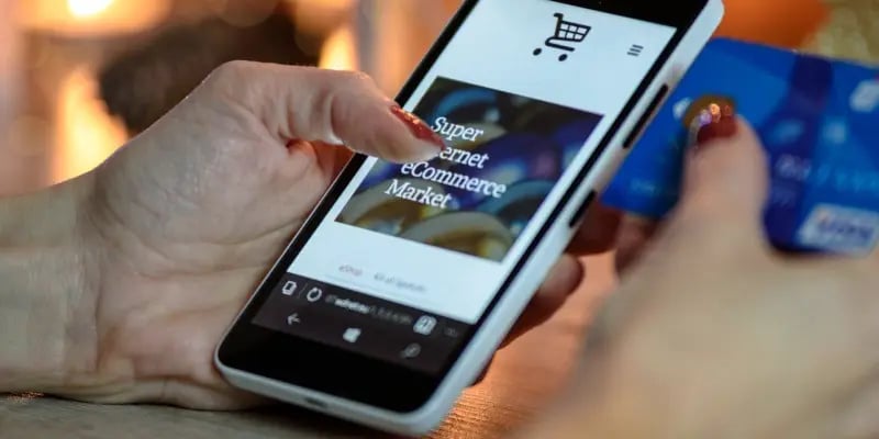 A person holds a smartphone displaying an online shopping site in one hand and a blue credit card in the other.