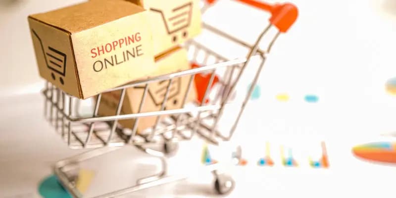 Miniature cardboard boxes labeled shopping online placed in a small shopping cart, with blurred charts and graphs in the background.