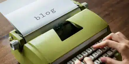 A person types on a green typewriter with a sheet of paper displaying the word blog.