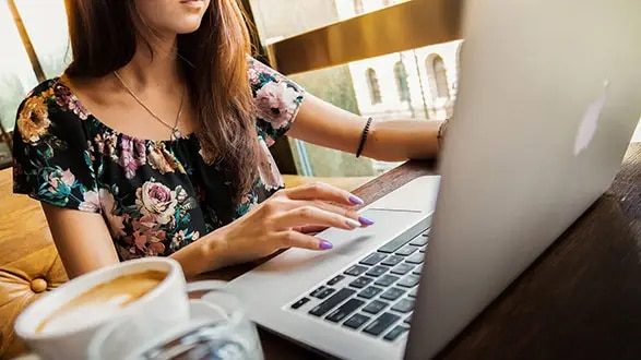 A person with long hair types on a laptop at a wooden table near a window. A cup of coffee sits nearby.