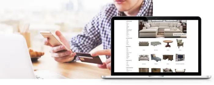 Person holding a credit card and smartphone near a laptop displaying an online furniture store with various furniture options visible on the screen.
